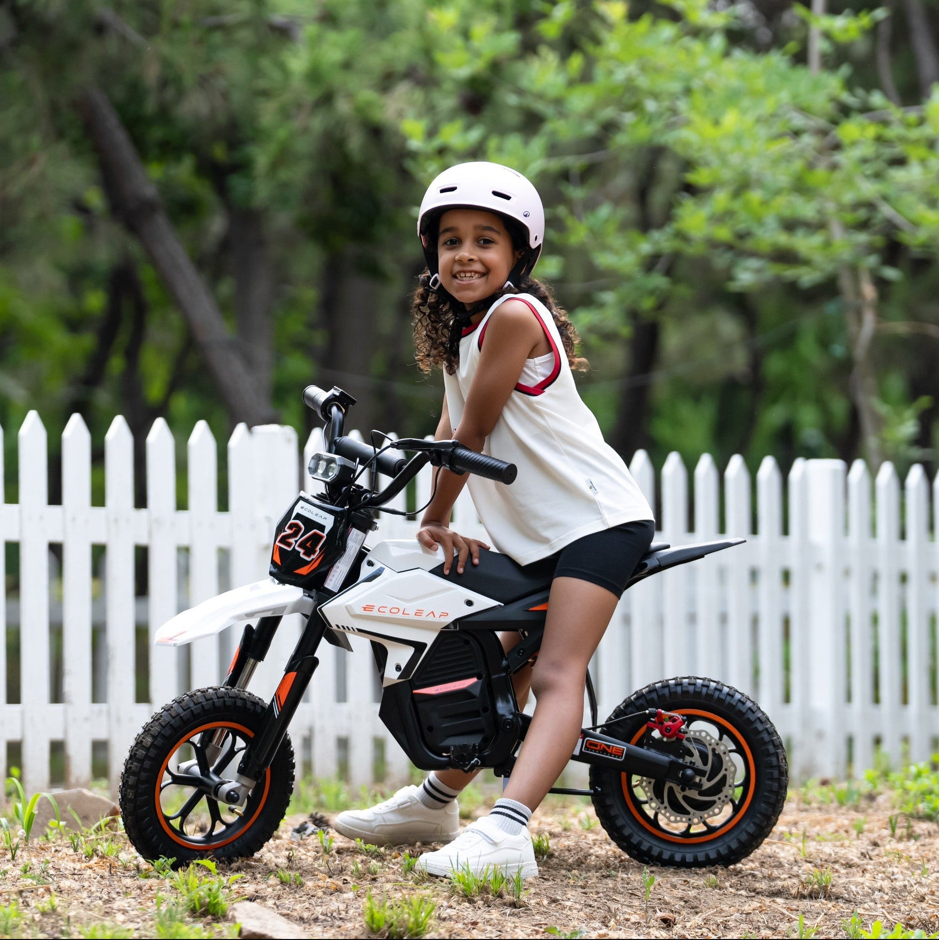 A young girl wearing a white helmet and a white sleeveless top with red trim sits on a small white and black electric dirt bike with orange accents. The scene is outdoors in front of a white picket fence with green trees in the background.