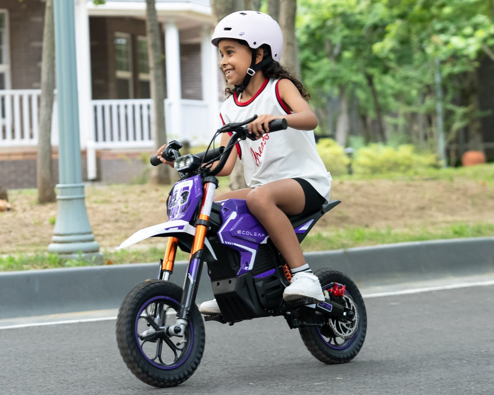 A young child wearing a white helmet rides a purple and orange electric mini dirt bike on an asphalt street, with trees and a house in the background.